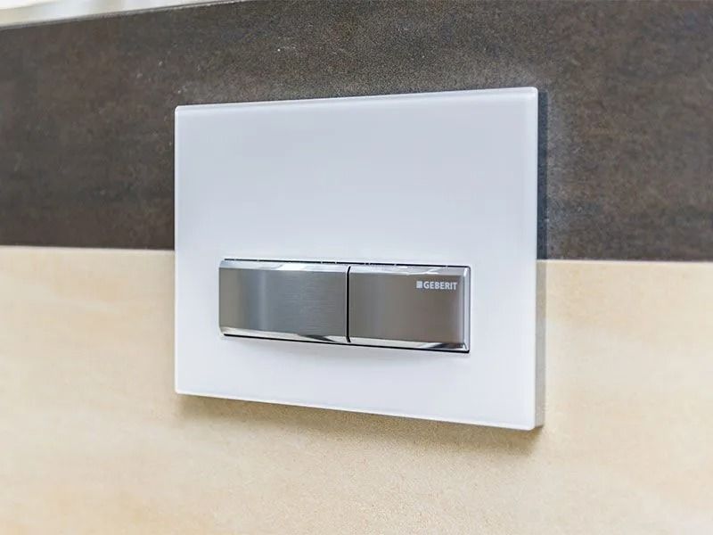 Weiße WC-Betätigungsplatte mit verchromter Zweimengenspültaste an gefliester Wand.
