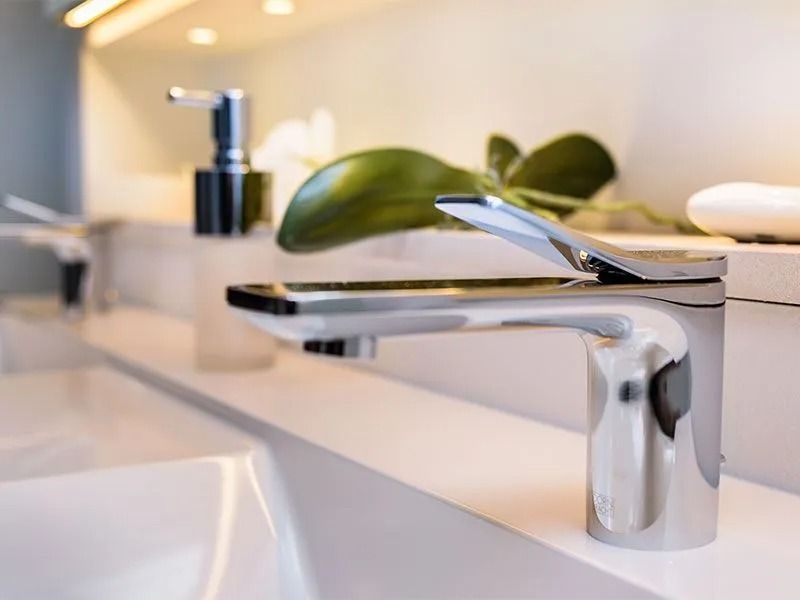 Moderne verchromte Waschtischarmatur auf einem Waschbecken mit Seifenspender und dekorativer Pflanze.
