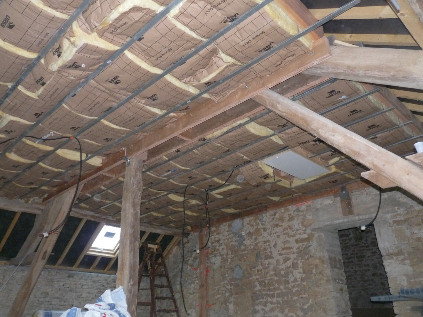 Grenier en rénovation avec charpente apparente, isolation, échelle et murs en pierre.