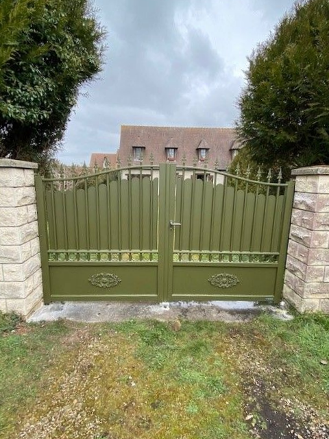Portail en métal vert avec piliers en pierre devant une maison.