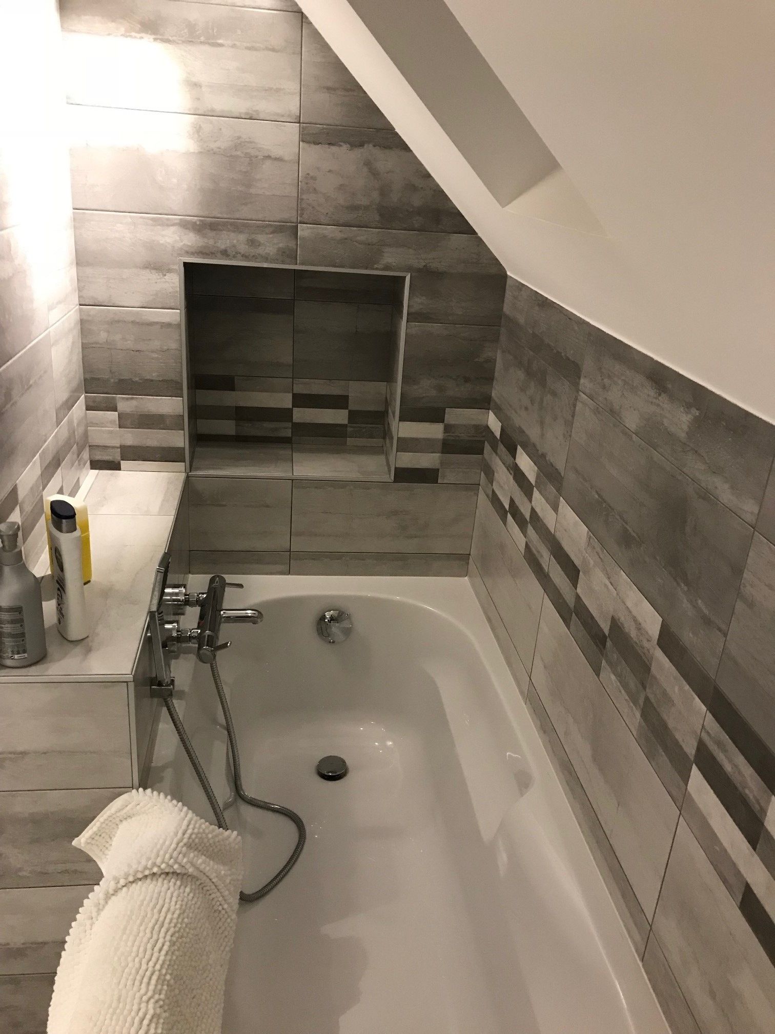 Salle de bains moderne avec baignoire blanche, douchette, murs carrelés gris et tapis sur le rebord.