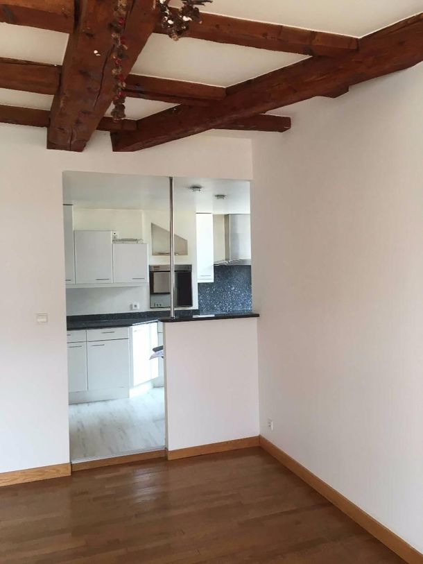 Pièce vide avec parquet et poutres apparentes au plafond, ouverte sur un coin cuisine blanc.