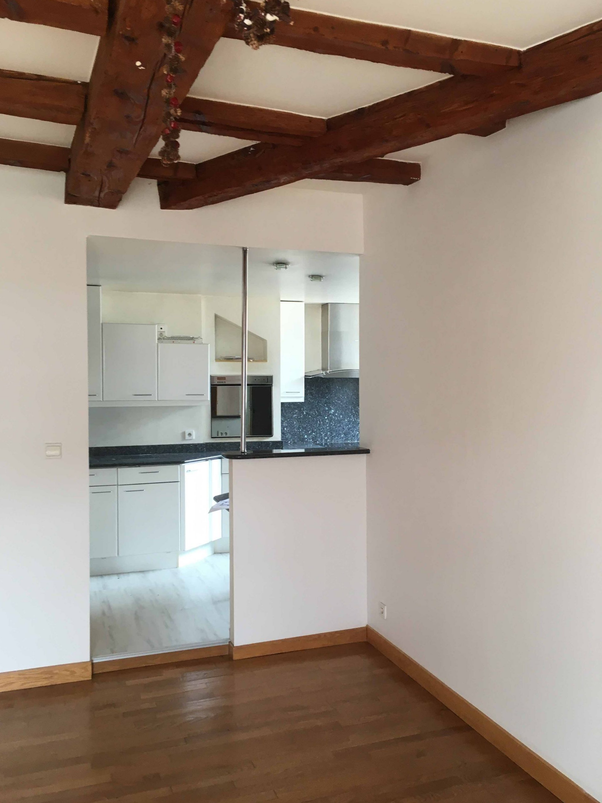 Pièce vide avec poutres apparentes, parquet et petite cuisine ouverte au fond.