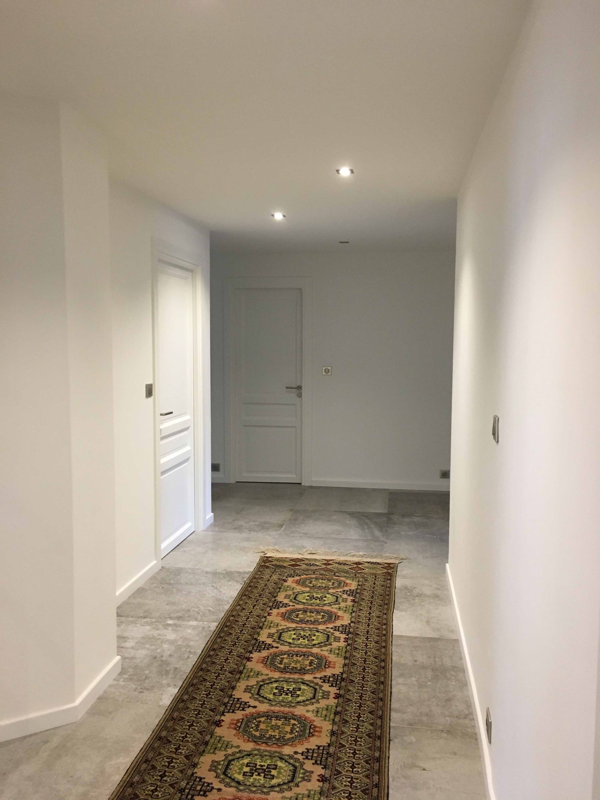 Couloir étroit aux murs blancs, carrelage et tapis à motifs menant à une porte fermée.