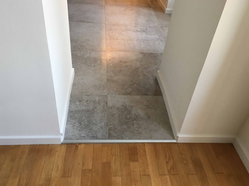 Couloir avec un sol carrelé gris menant à un seuil en parquet.