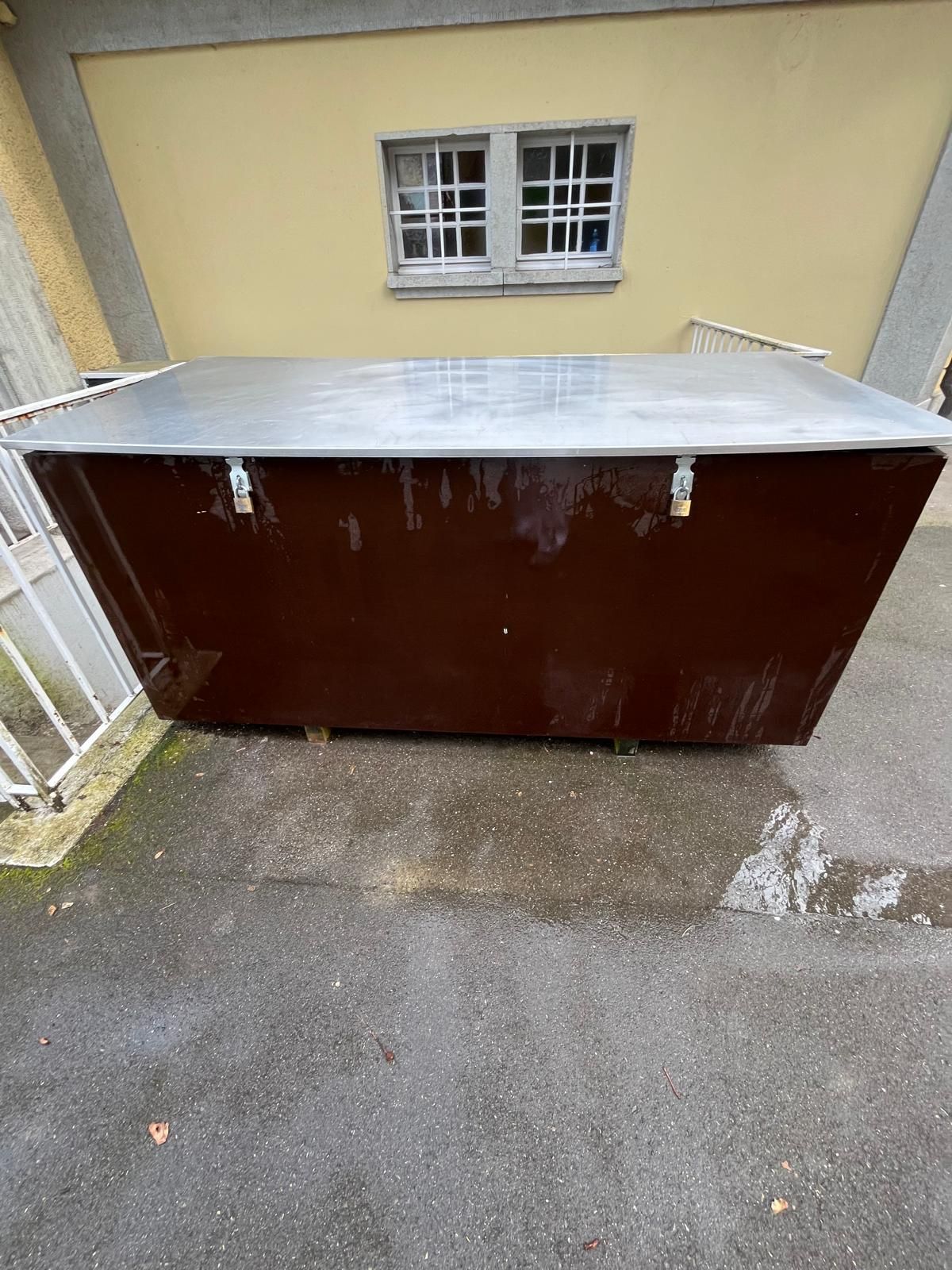 Vor einem Gebäude steht ein brauner Müllcontainer mit Metalldach.