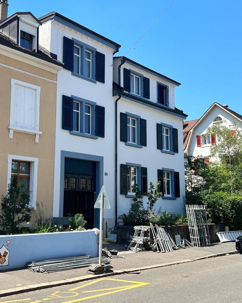 Ein weißes Gebäude mit schwarzen Fensterläden steht neben einem hellbraunen Gebäude