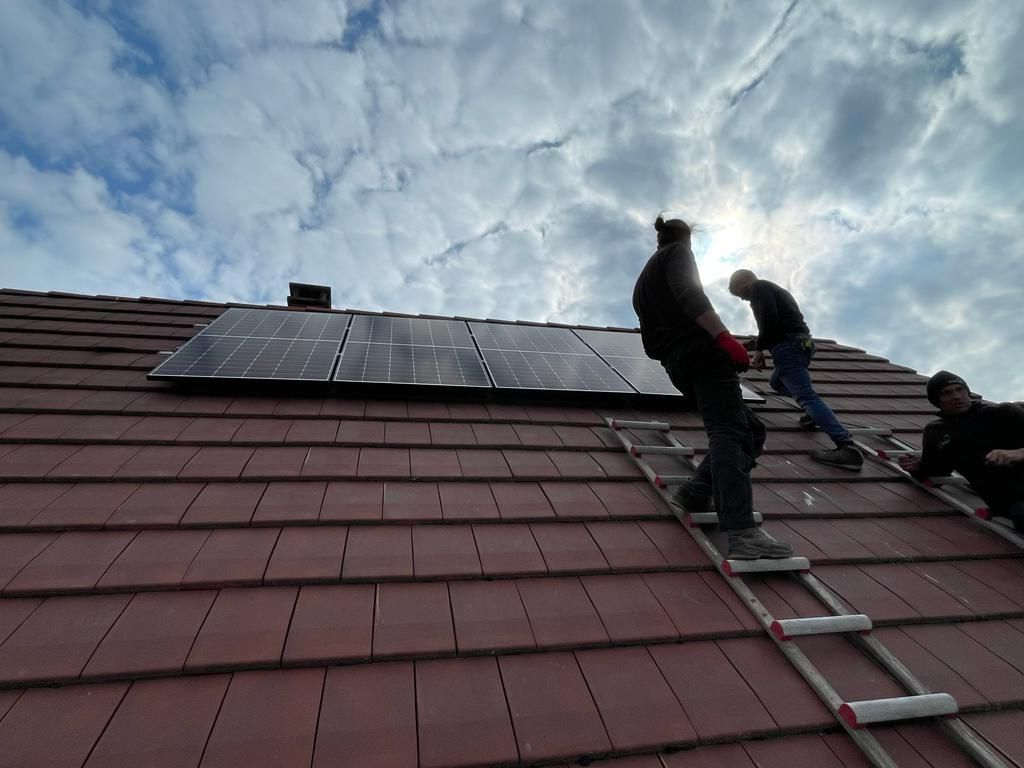 Des ouvriers installent des panneaux solaires sur un toit de tuiles rouges, grimpant à une échelle.