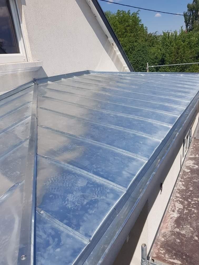 Toit en métal argenté à nervures linéaires, contrastant avec un mur blanc et un ciel bleu.