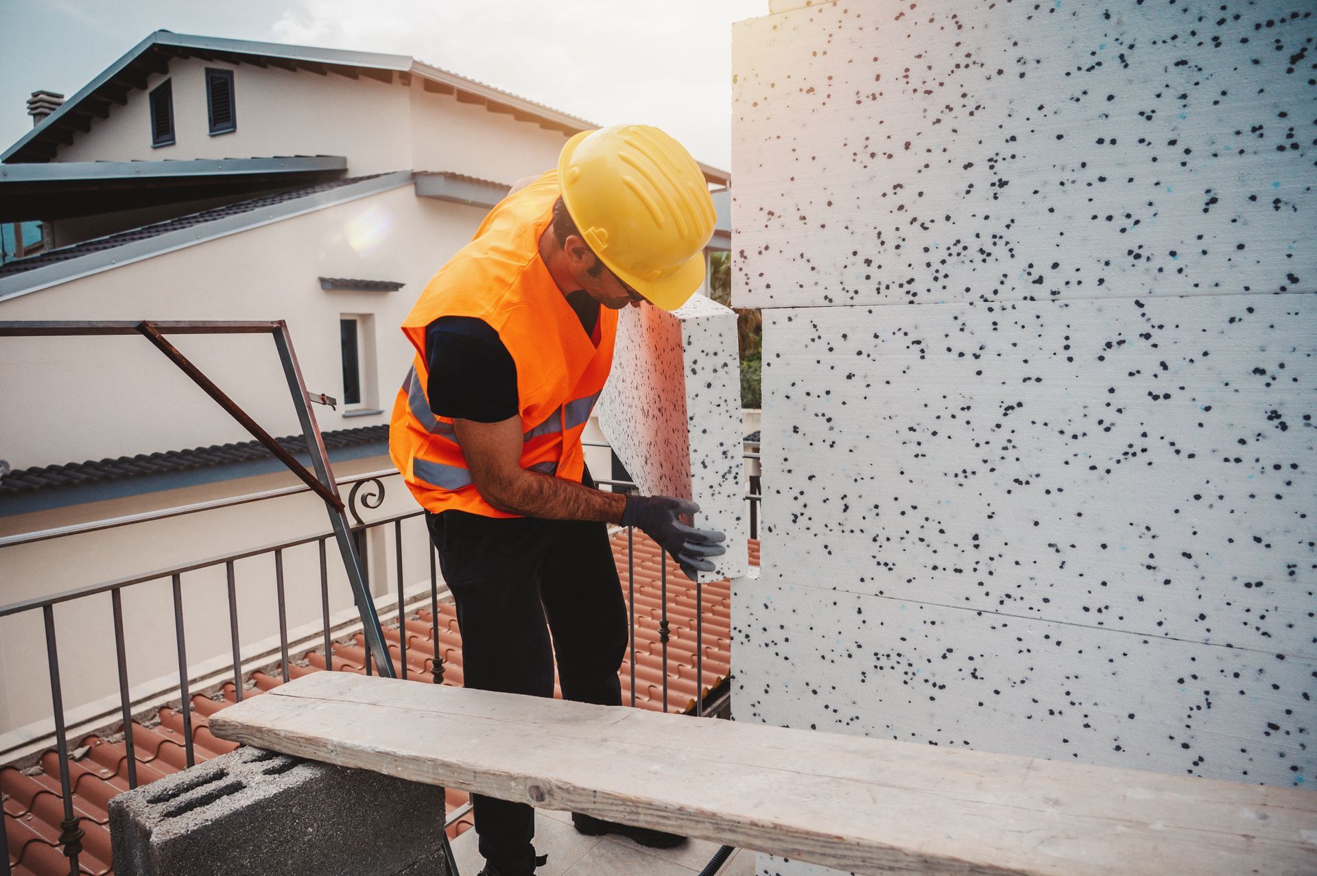 Un ouvrier du bâtiment installe de l'isolant sur un mur extérieur, portant un casque et un gilet de sécurité.