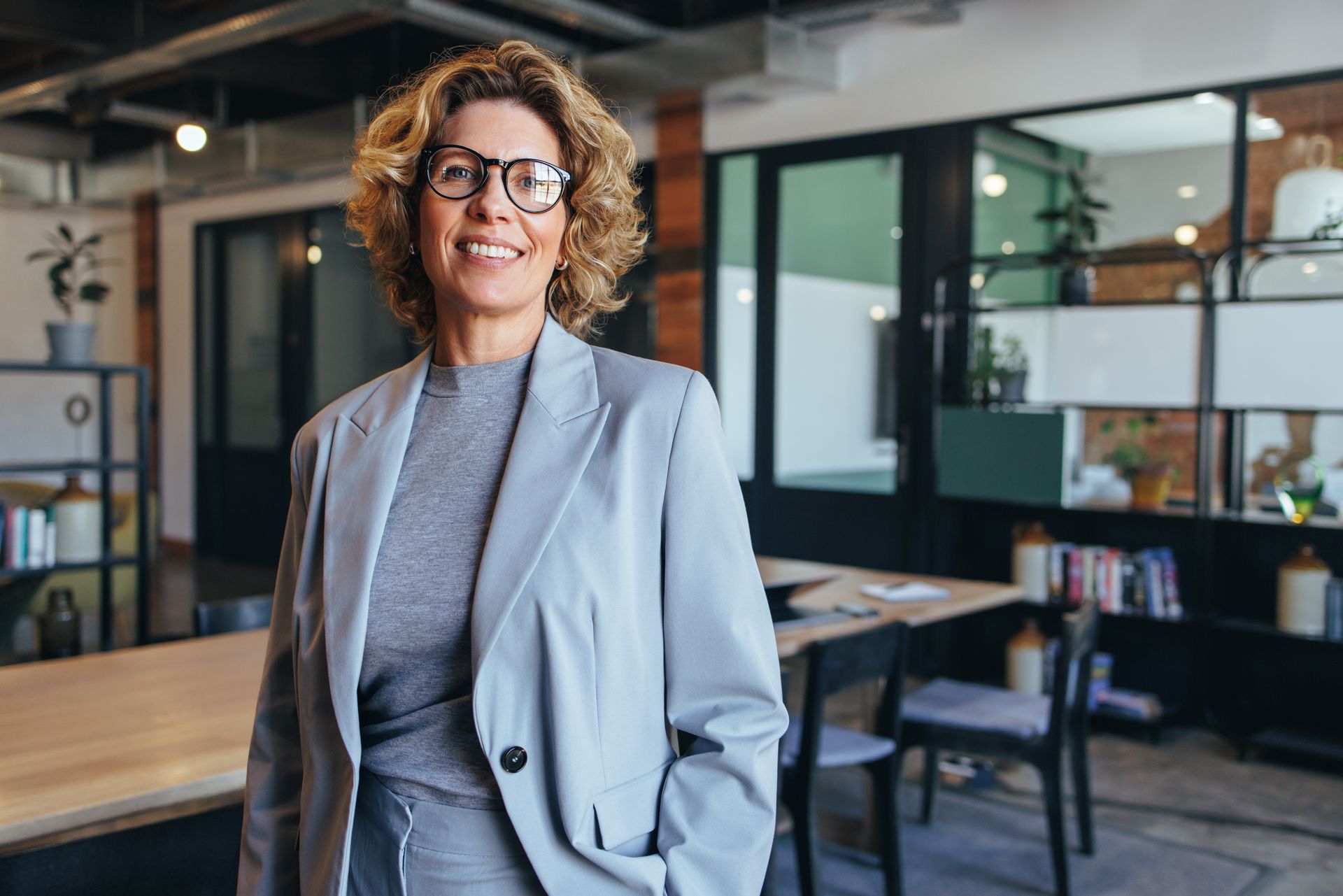 Une femme en blazer gris et lunettes sourit dans un bureau.