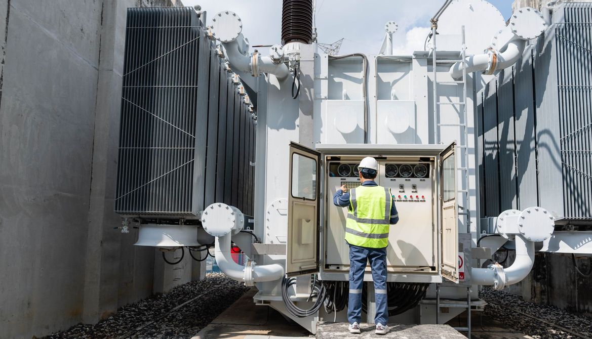 Ein Arbeiter in Sicherheitsweste inspiziert im Freien zwischen grossen Versorgungsanlagen einen Container für elektrische Ausrüstung.