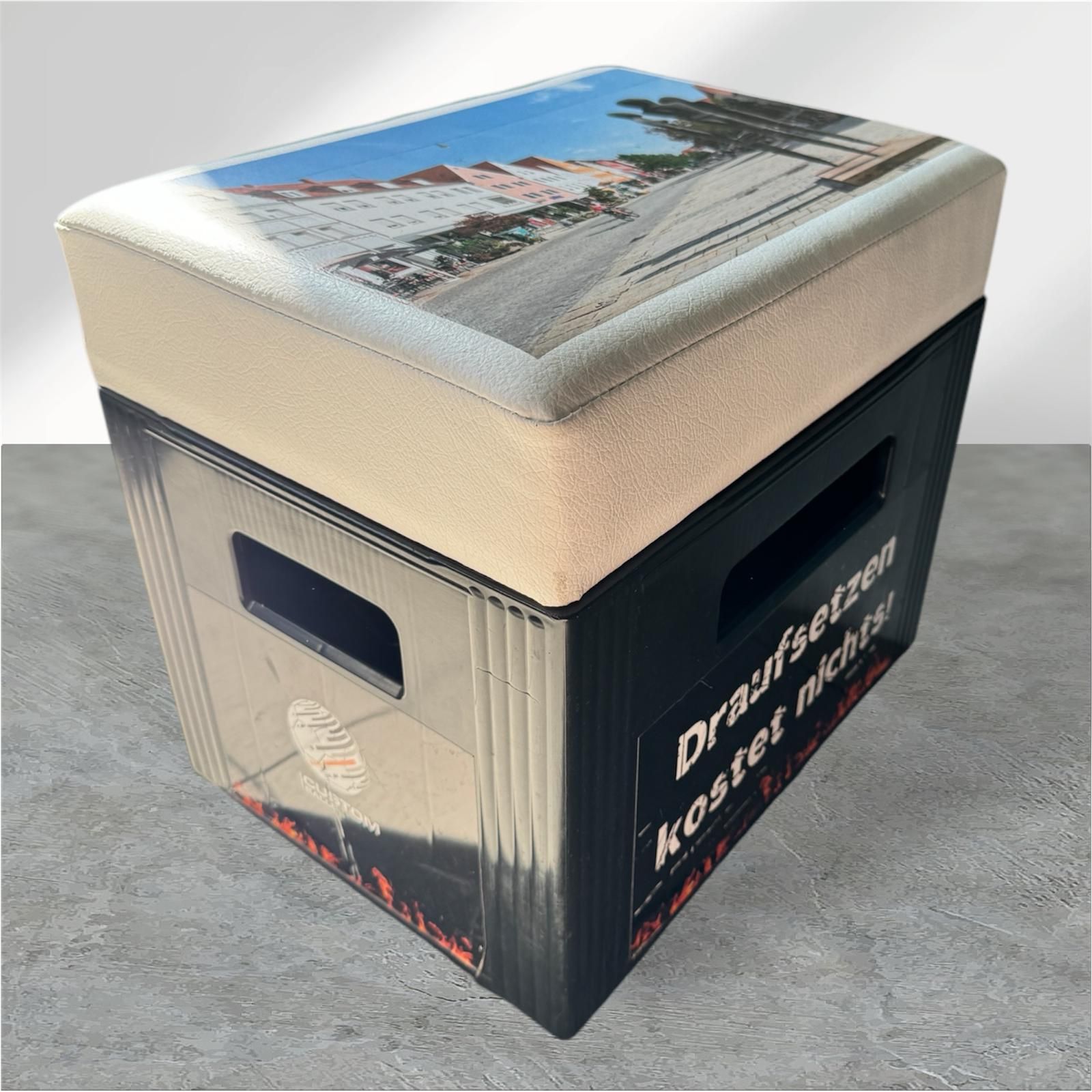 Schwarze Kiste mit gepolstertem Deckel. Das Kissen ist mit einer Straßenszene bedruckt.