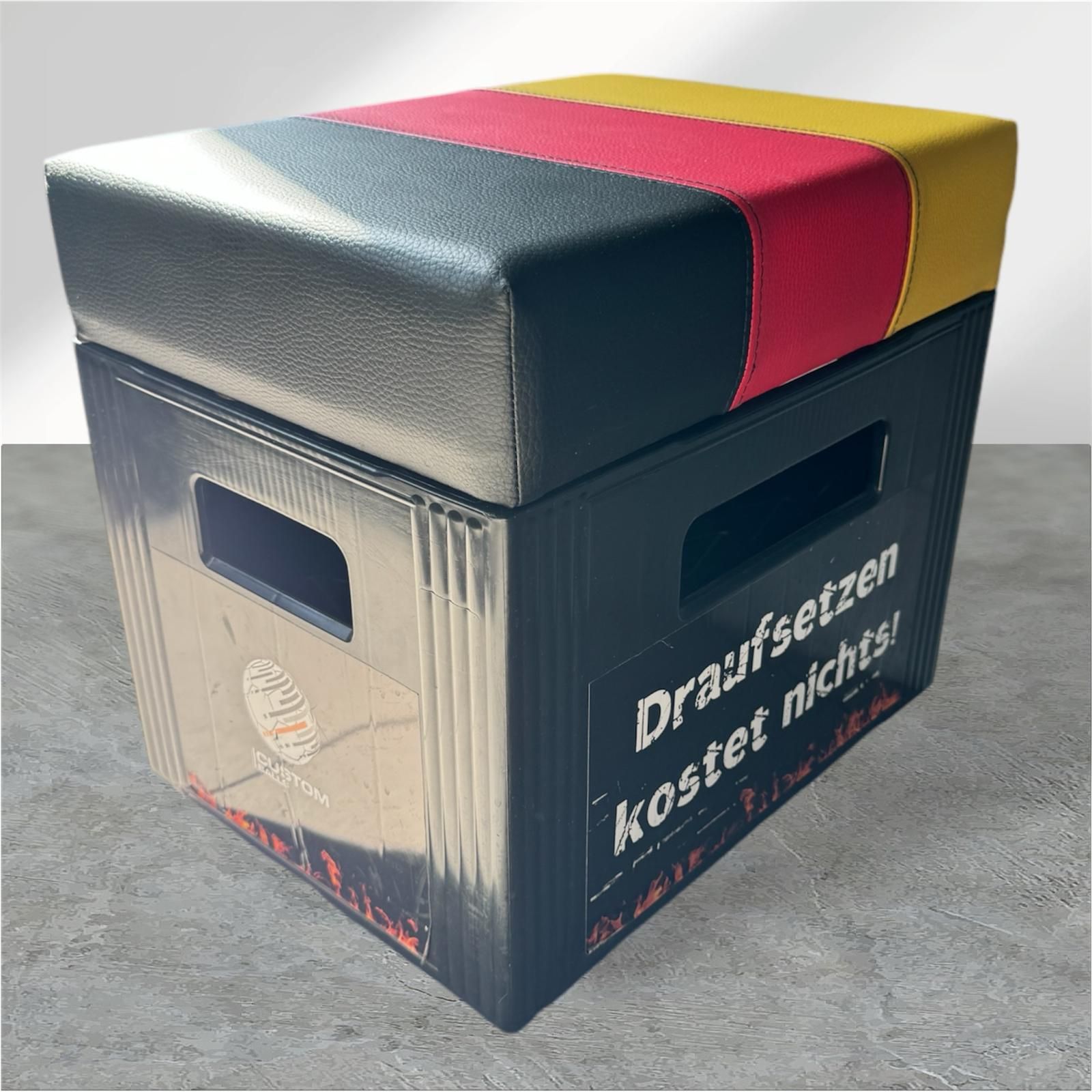 Schwarze Kiste, umfunktioniert als Hocker, mit gepolstertem Deckel in den Farben der deutschen Flagge.
