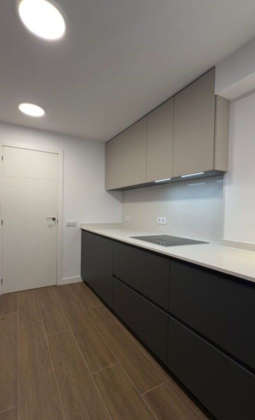 Cocina moderna con paredes blancas, suelo de madera oscura, armarios bajos negros, encimeras claras y electrodomésticos empotrados.
