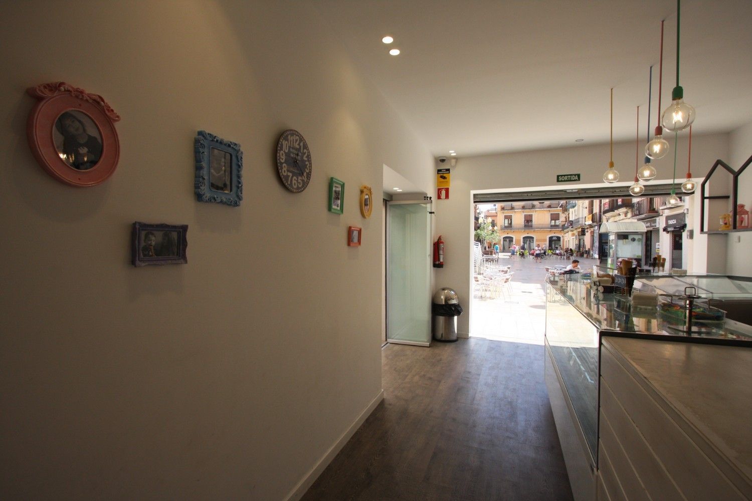 Un estrecho pasillo de cafetería con obras de arte en las paredes conduce a una barra luminosa y una zona de asientos al fondo.
