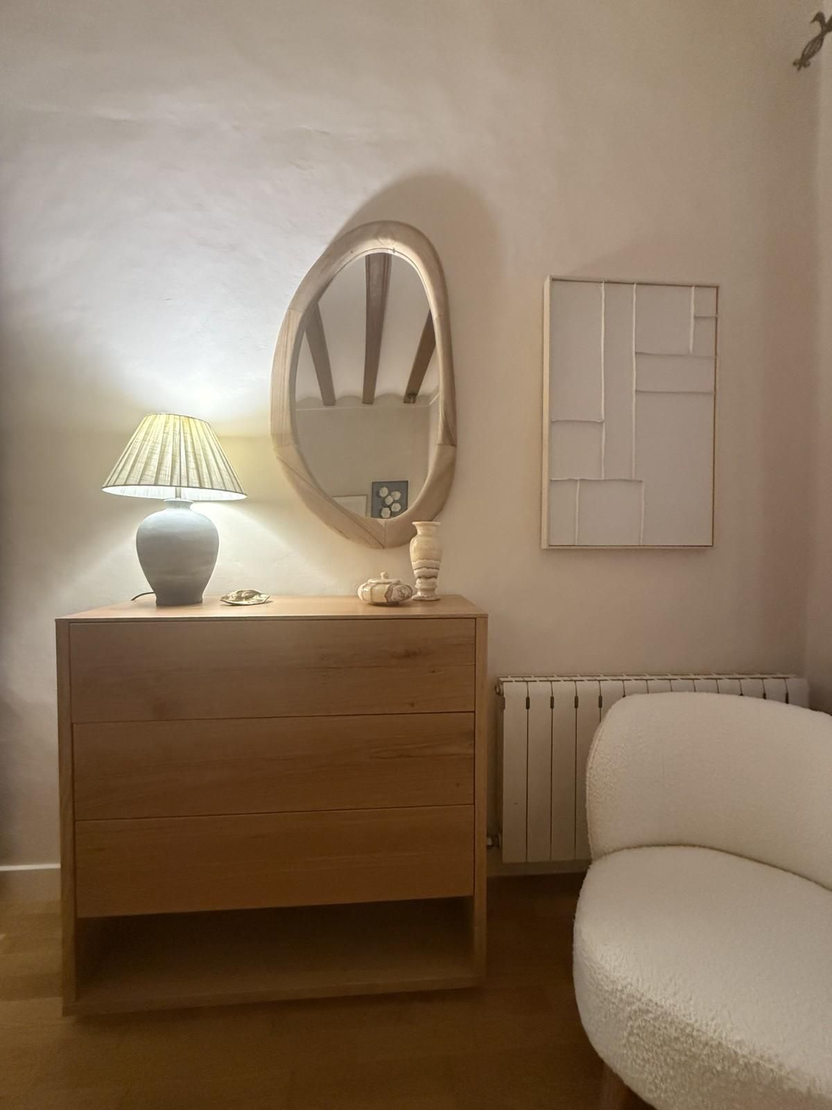 Acogedor rincón de dormitorio con lámpara encendida sobre una cómoda de madera, espejo ovalado, cuadro en la pared, radiador y silla blanca.