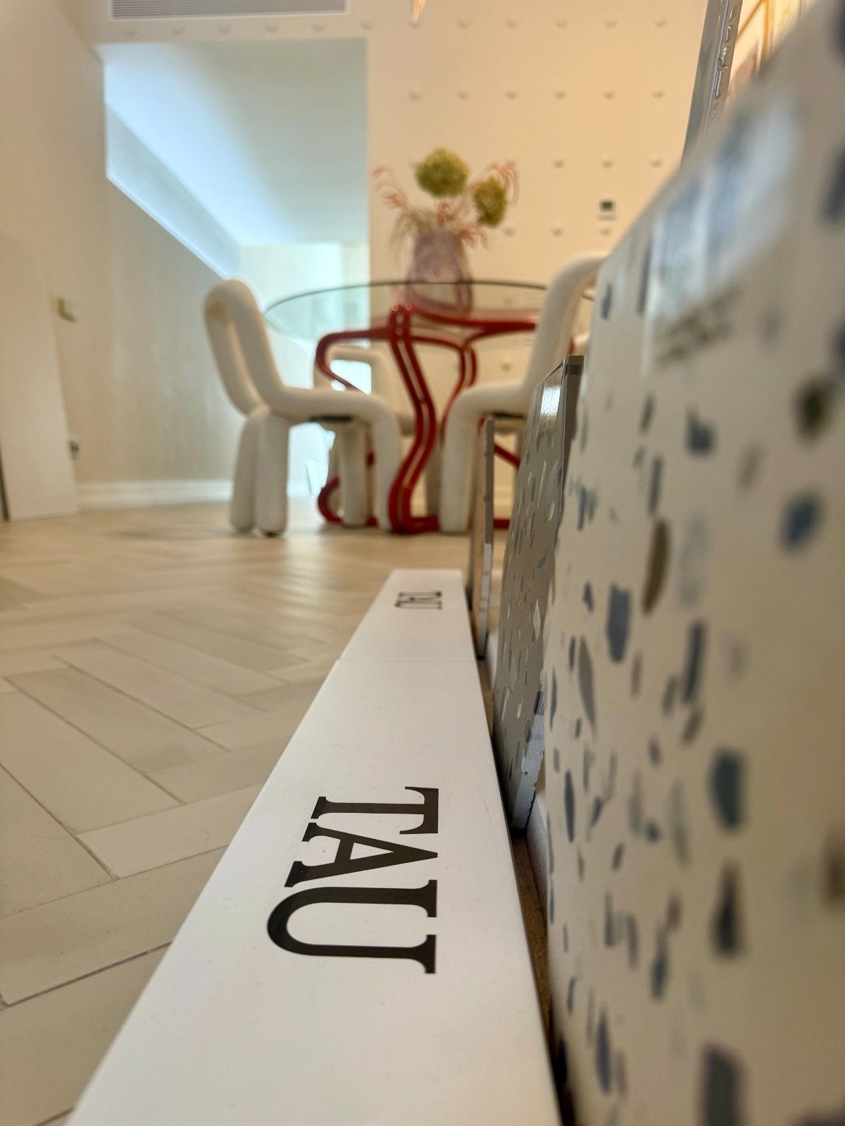 Logotipo de TAU en una pizarra blanca en una habitación luminosa con sillas blancas y una mesa roja.