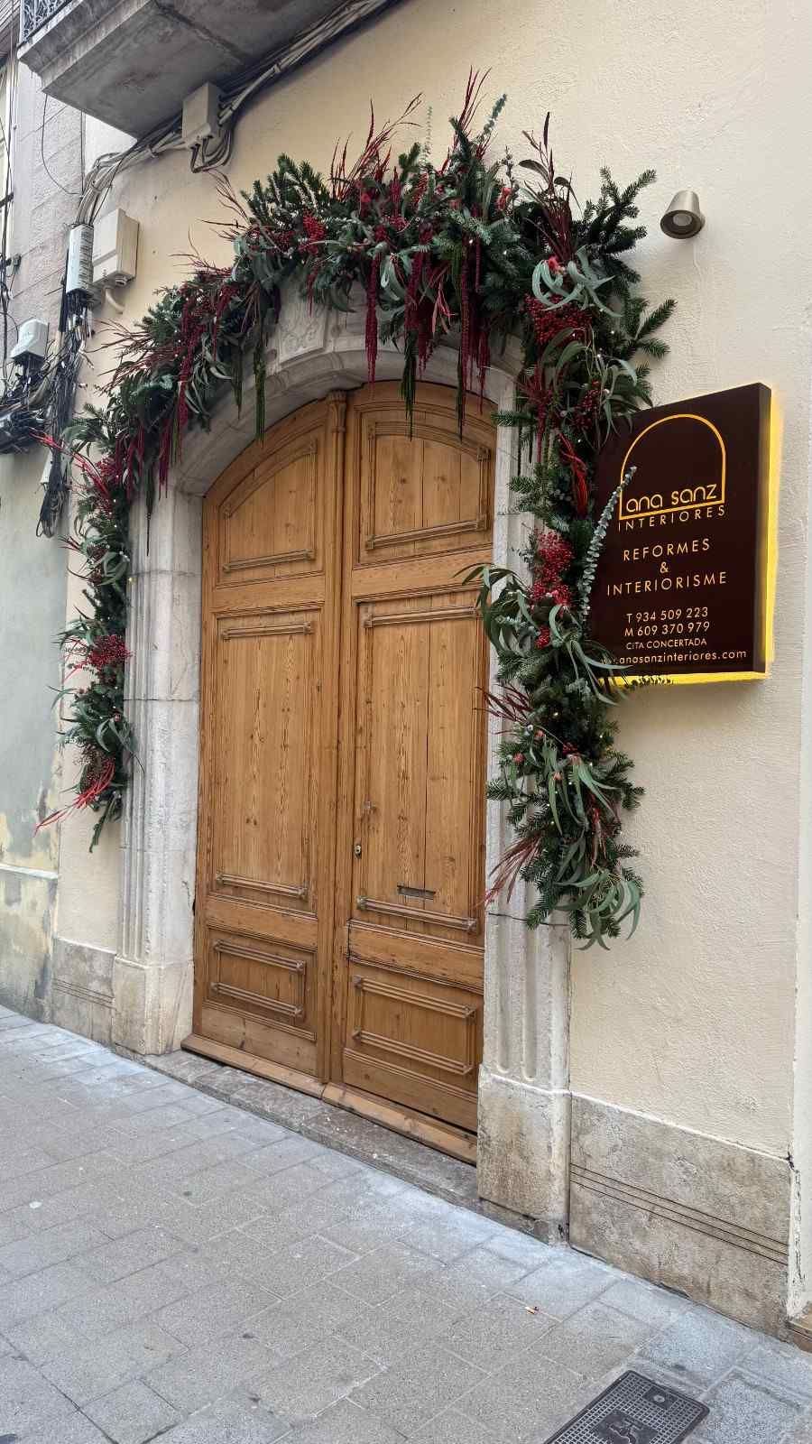 Puerta de iglesia de madera decorada con guirnalda verde y flores rojas junto a una placa en la pared.