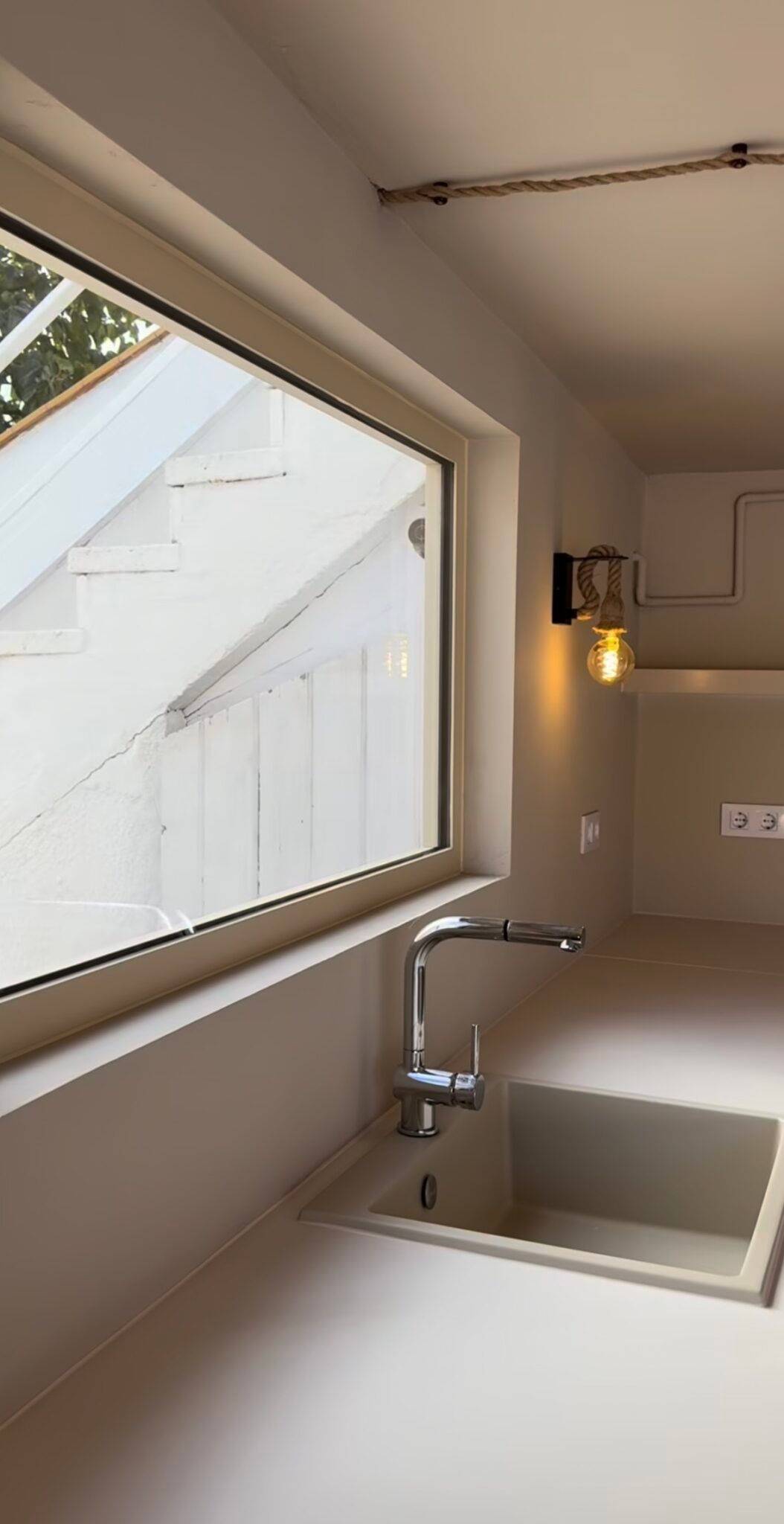 Cocina pequeña con una ventana sobre un fregadero y grifo de acero inoxidable, encimeras de color beige y una lámpara de pared.