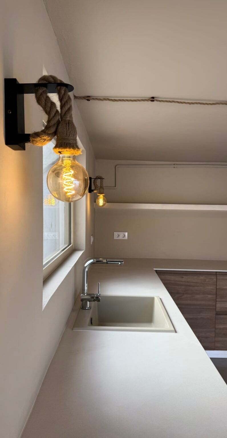Cocina moderna con fregadero, encimera, ventana y lámparas colgantes sobre una pared de color beige.