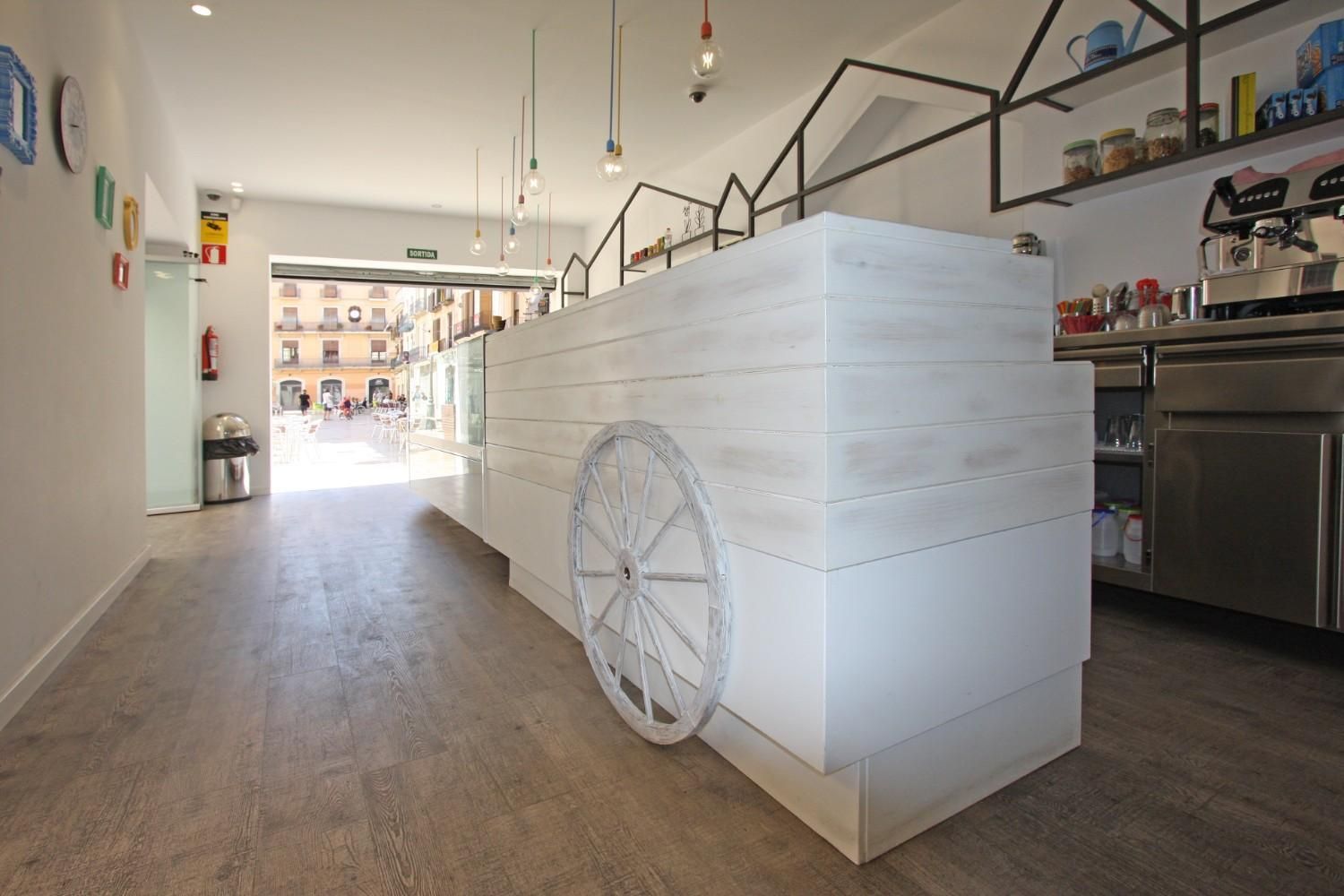 Cafetería luminosa con una larga barra blanca y una gran rueda como elemento decorativo en un interior moderno.