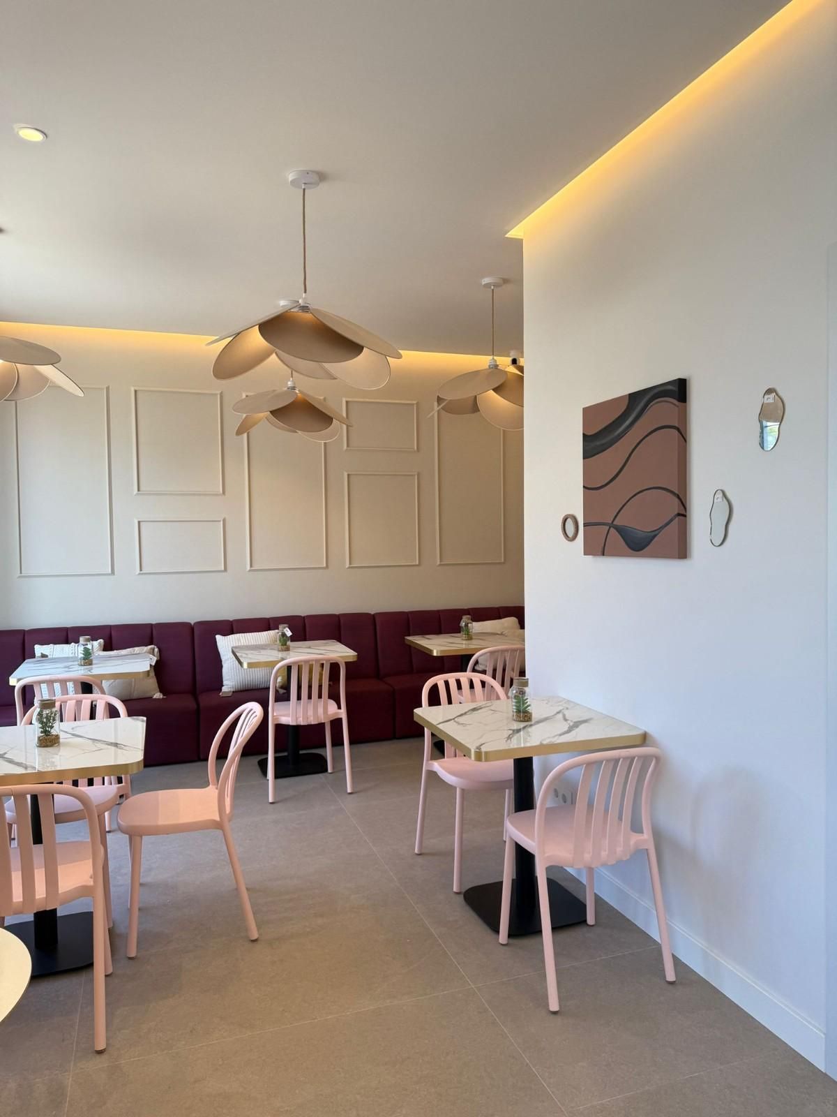 Interior minimalista de una cafetería con sillas blancas, mesas pequeñas, bancos color burdeos y lámparas colgantes de luz cálida.