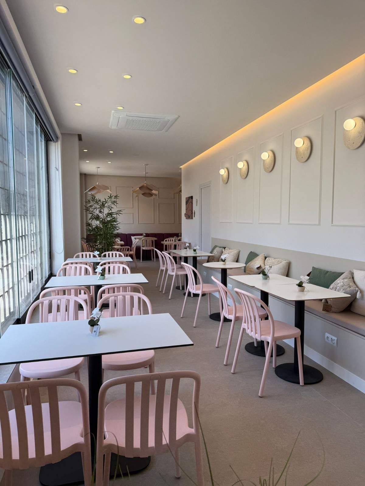 Interior luminoso de la cafetería con sillas rosas, mesas blancas, apliques de pared y grandes ventanales a lo largo de un lado.