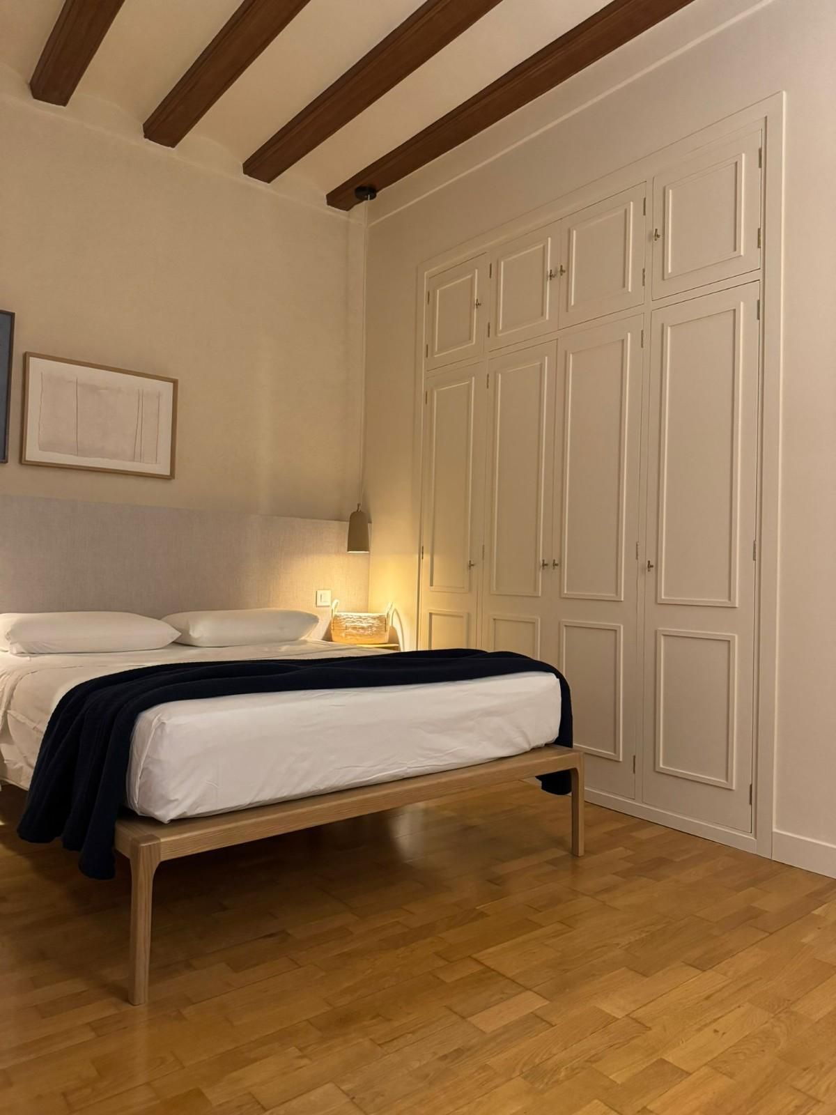 Dormitorio minimalista con cama, puertas de armario blancas, vigas vistas y una lámpara de noche cálida.