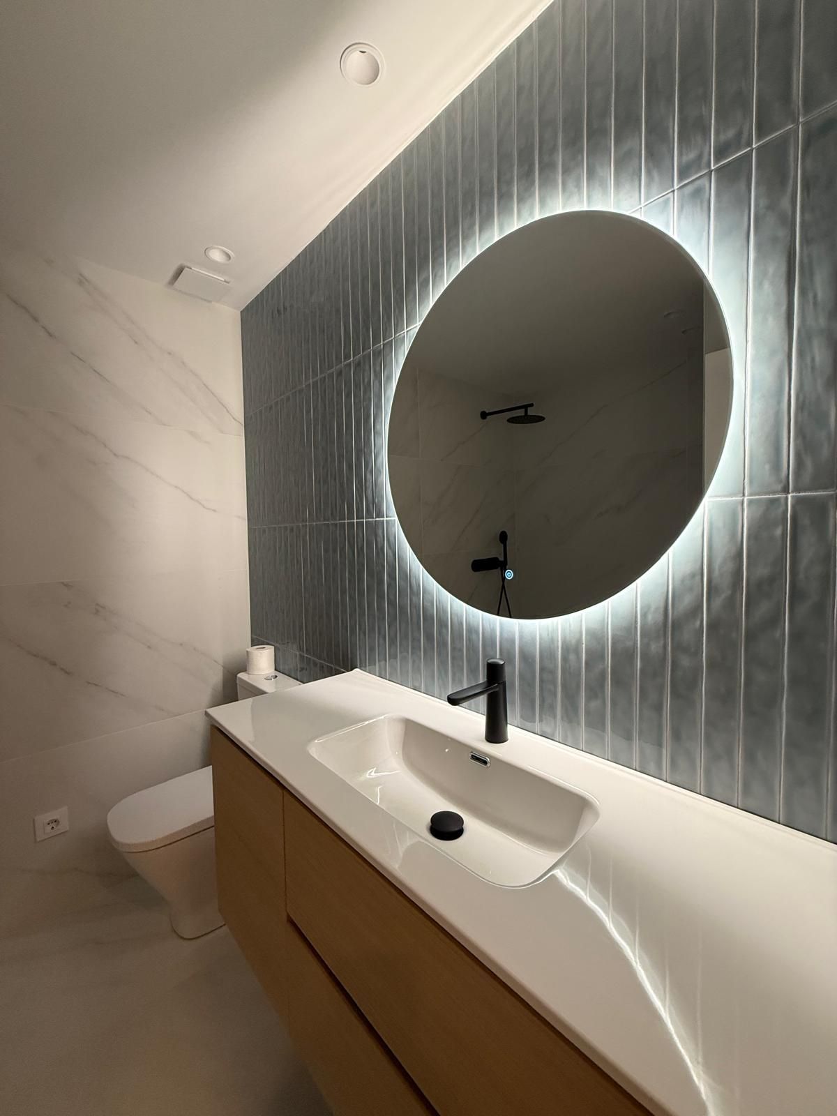 Tocador de baño moderno con espejo redondo retroiluminado, lavabo blanco y armarios de madera.