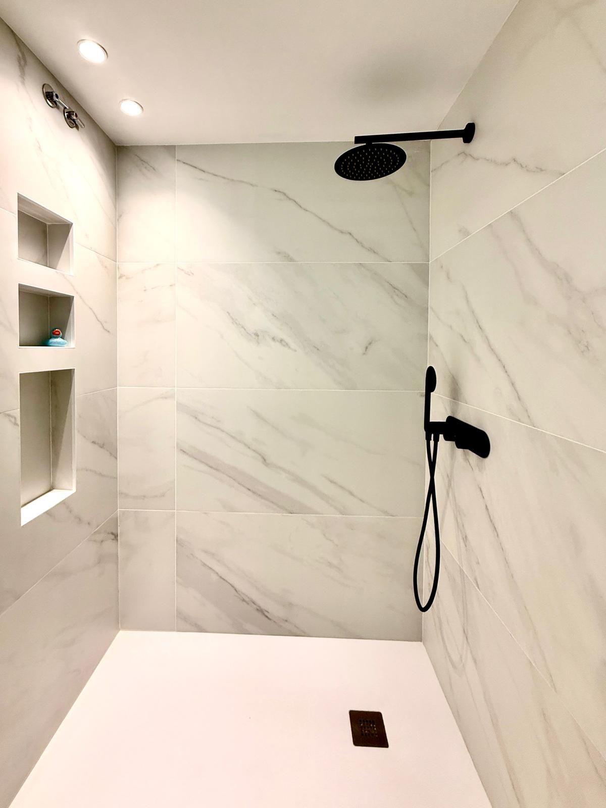 Ducha moderna revestida de mármol blanco con grifería negra y estantes integrados.