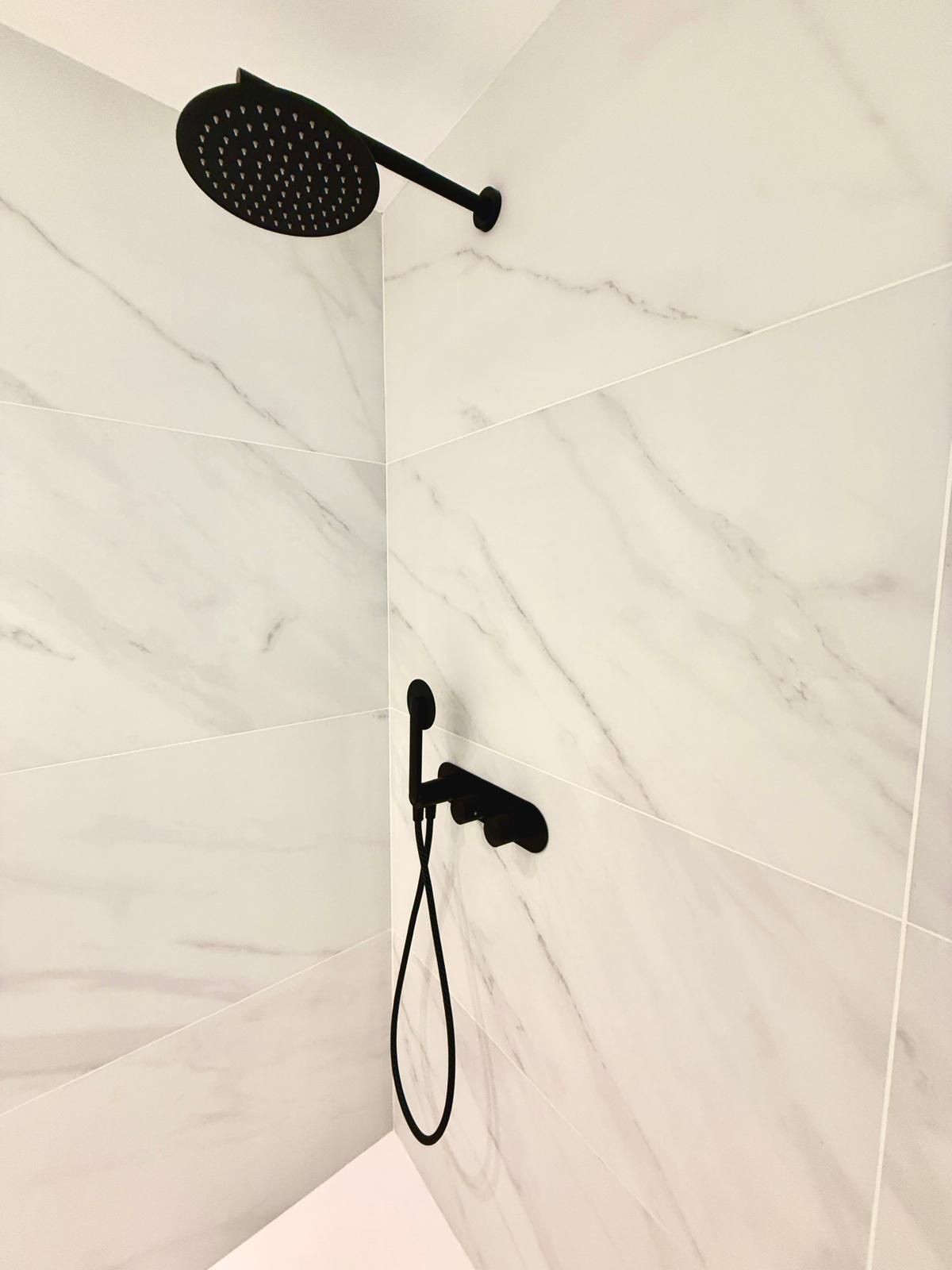 Cabezal de ducha negro y rociador de mano sobre una pared de ducha alicatada en blanco.