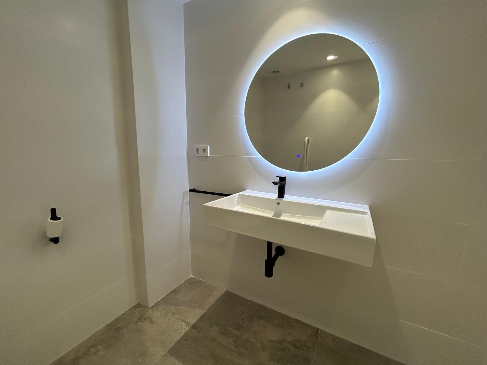 Baño moderno con azulejos blancos, lavabo suspendido y espejo redondo iluminado.