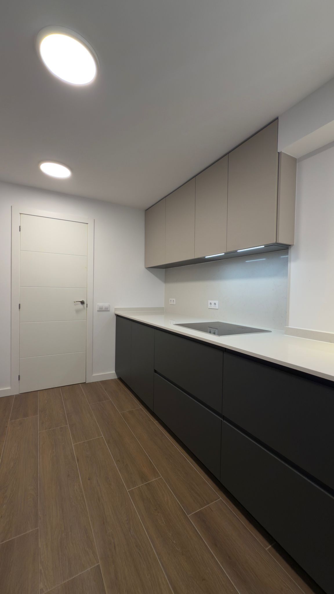Cocina moderna y estrecha con armarios oscuros, encimeras blancas, suelo de madera y puerta cerrada al final.