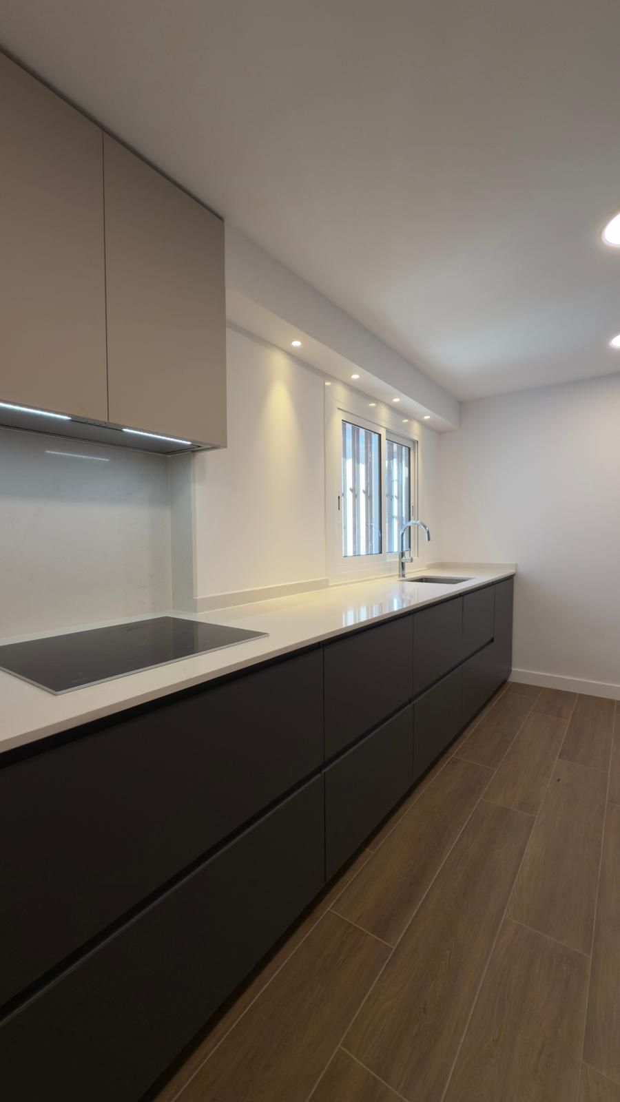 Cocina moderna con armarios bajos oscuros, encimeras claras, suelo de madera y luz natural en la ventana.