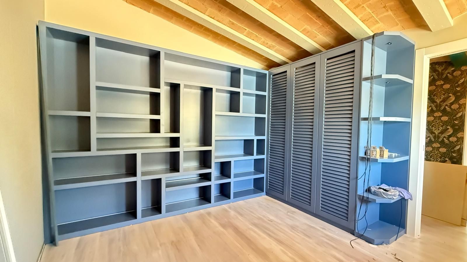 Pared con estanterías empotradas de color gris en una habitación luminosa con suelos de madera y vigas de techo a la vista.