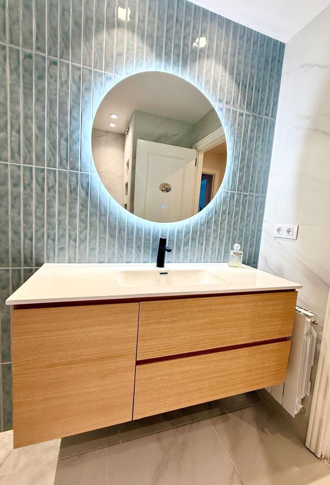Tocador de baño moderno con gabinetes de madera, grifo negro y espejo redondo sobre una pared de azulejos azules.