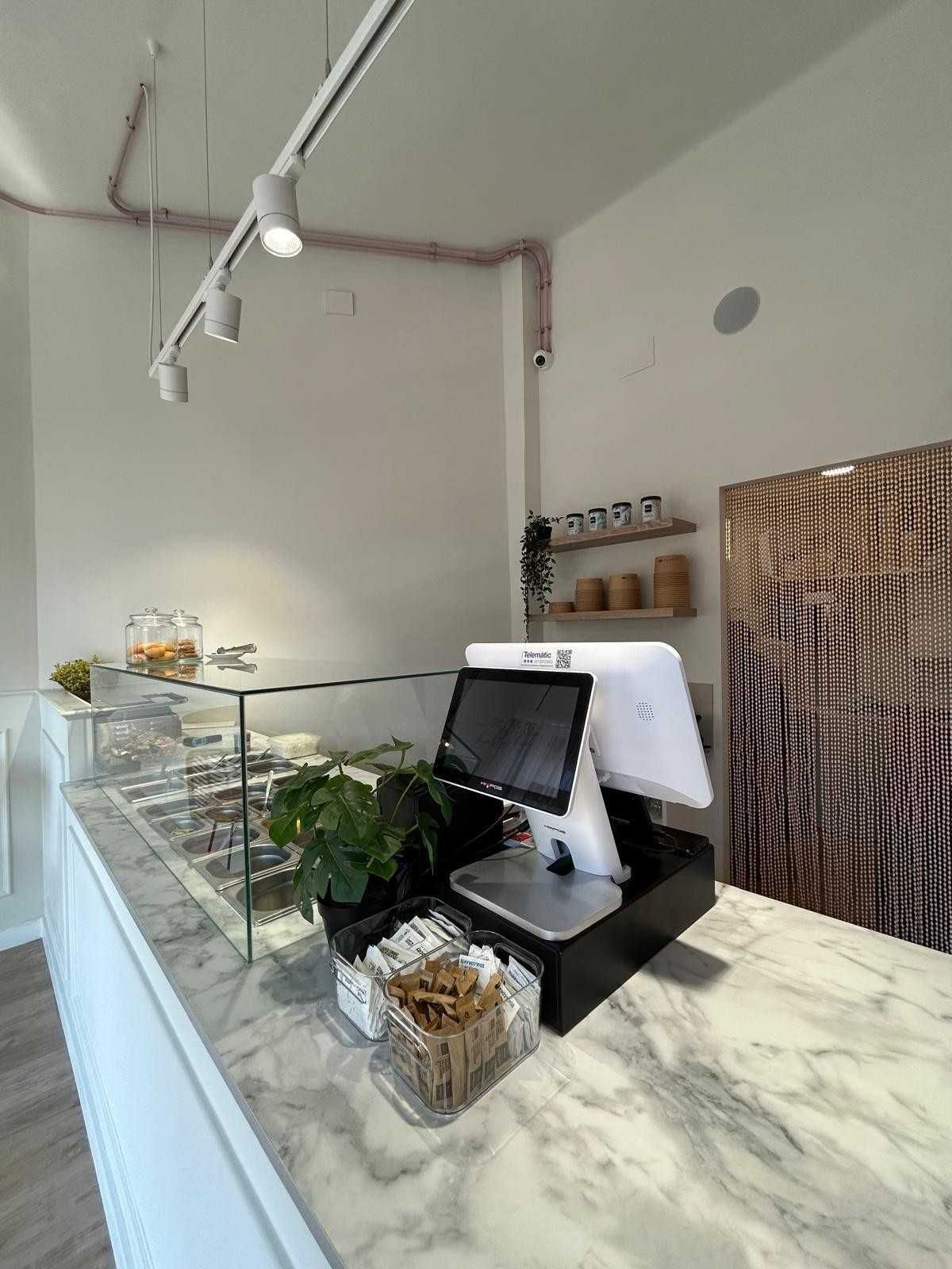 Barra de cafetería moderna con vitrina de cristal, encimera de mármol, plantas y pizarra blanca con el menú.