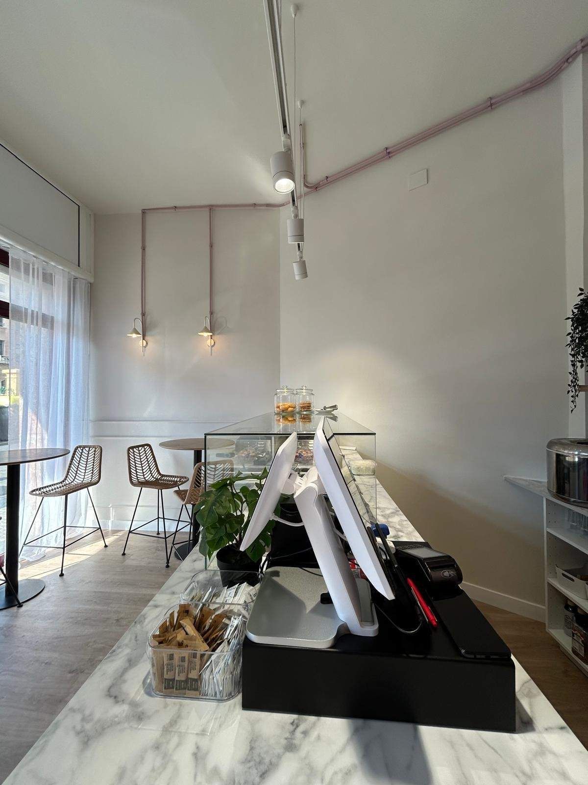 Interior de cafetería de color blanco brillante con una larga mesa repleta de plantas y elementos decorativos cerca de grandes ventanales.