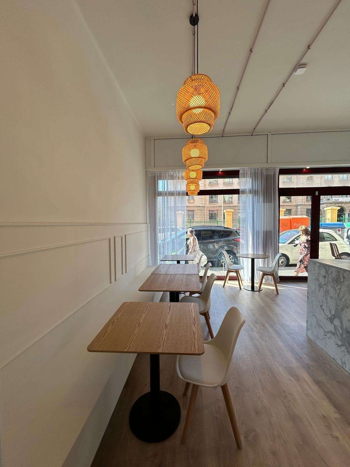 Interior de la cafetería con mesas de madera, lámparas colgantes y luz solar que entra por las ventanas delanteras.