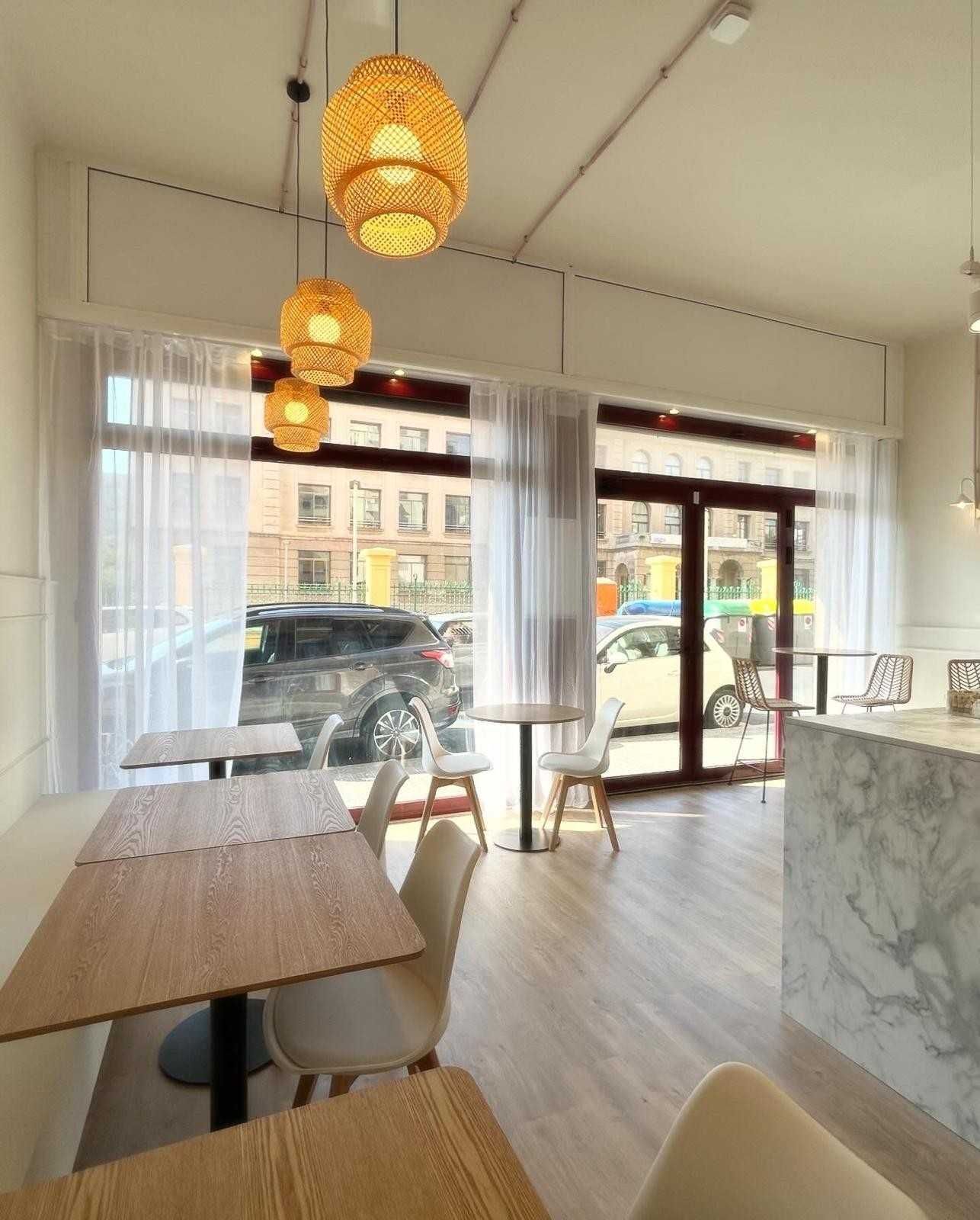 Interior luminoso y moderno de una cafetería con lámparas colgantes de mimbre, mesas y grandes ventanales con vistas a la calle.