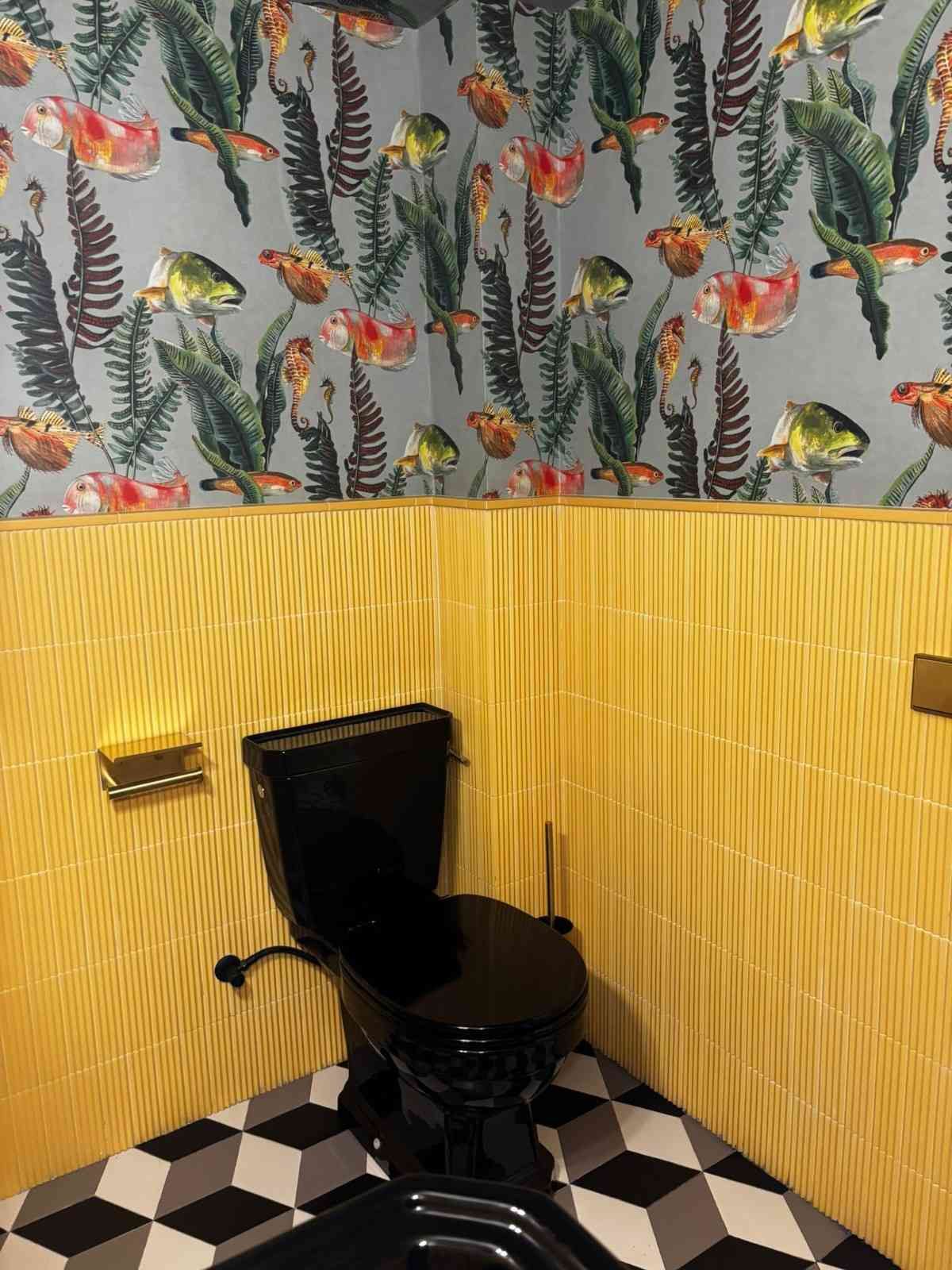 Baño pequeño con inodoro negro, paredes amarillas texturizadas, papel pintado con estampado de hojas de plátano y suelo de baldosas a cuadros blancas y negras.