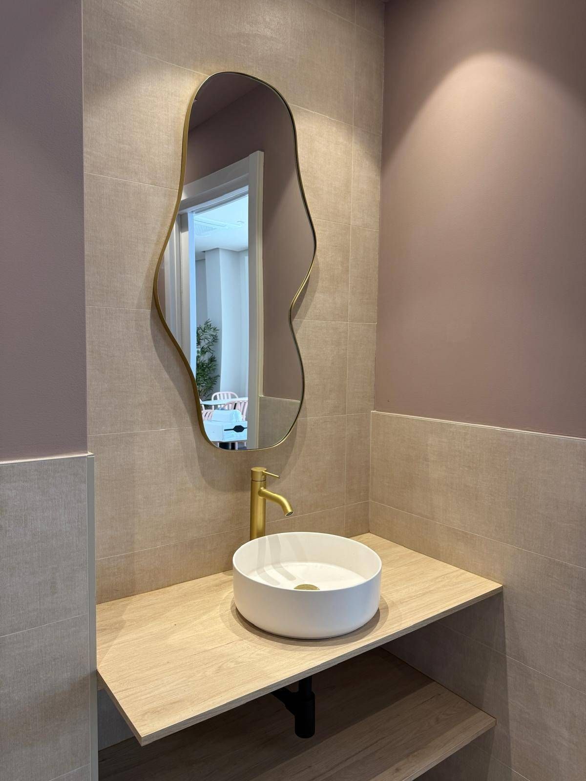 Tocador de baño moderno con lavabo redondo blanco, grifo dorado, espejo ondulado y encimera beige.