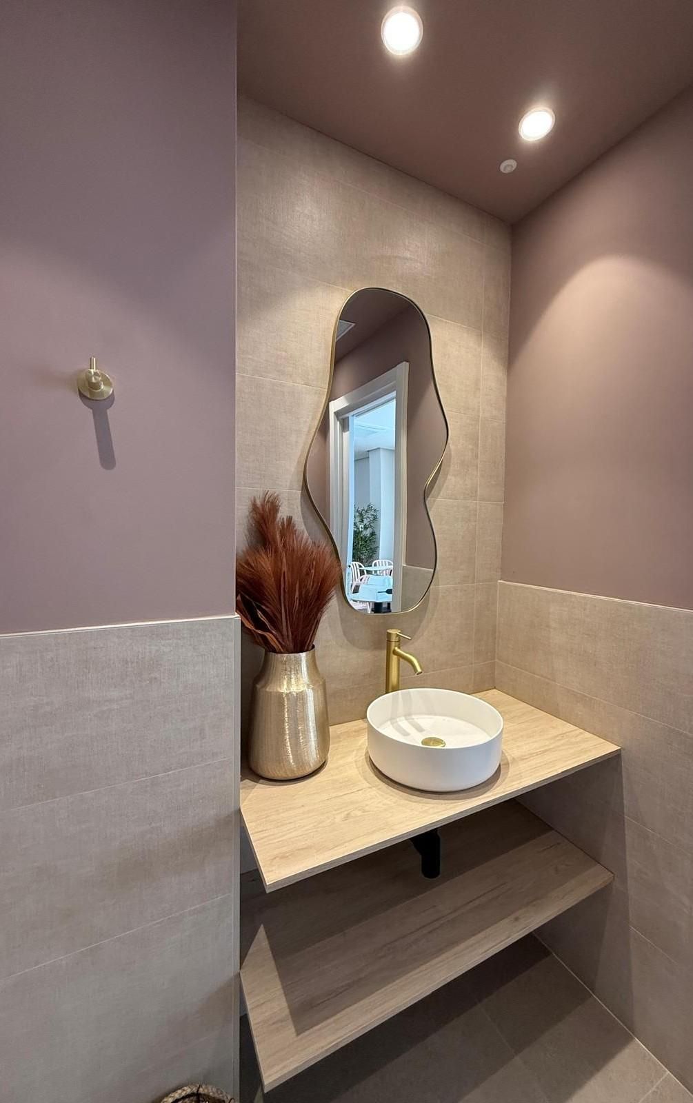 Pequeño rincón de tocador con paredes color malva, espejo, lavabo sobre encimera, jarrón y estantes bajo luces cálidas.