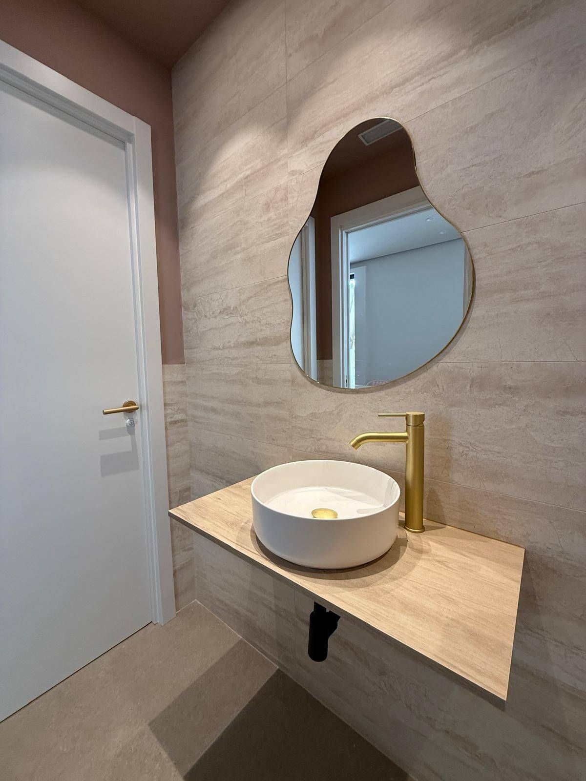 Tocador de baño moderno con espejo redondo, grifo dorado y lavabo sobre encimera, junto a una puerta blanca.