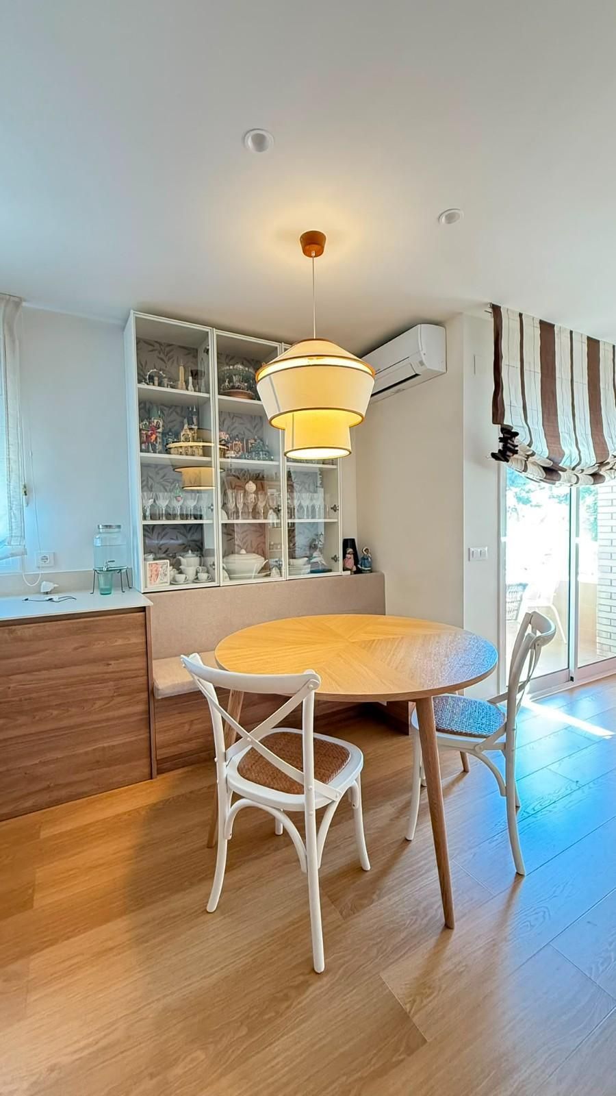 Comedor con mesa redonda de madera, sillas blancas, vitrinas y una lámpara colgante cerca de grandes ventanales.