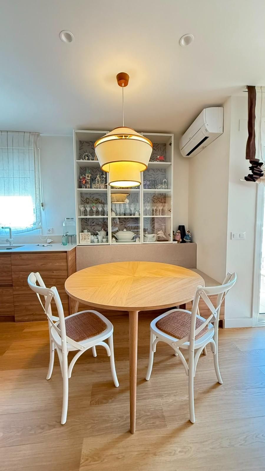 Comedor luminoso con mesa redonda de madera, sillas blancas, lámpara colgante, armarios y aire acondicionado mural.