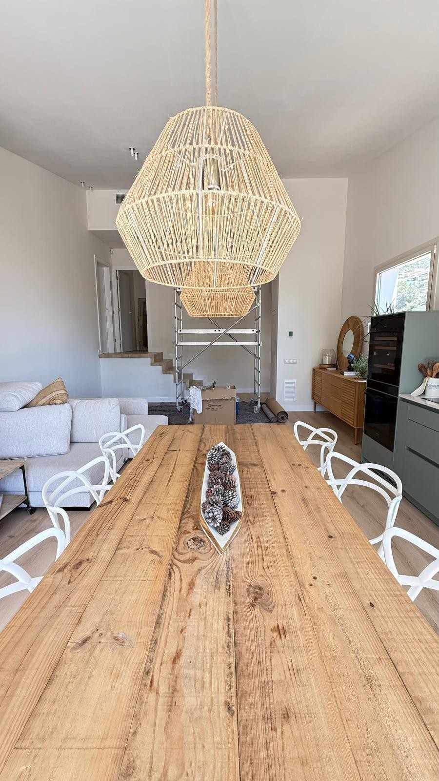 Mesa de comedor larga de madera con sillas blancas y lámparas colgantes tejidas en un espacio abierto y luminoso.