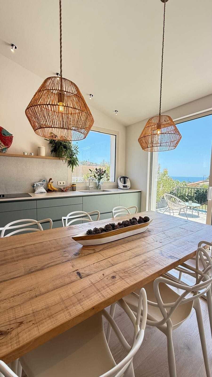 Luminosa cocina-comedor con mesa de madera, lámparas colgantes tejidas y vistas al océano a través de las puertas abiertas.
