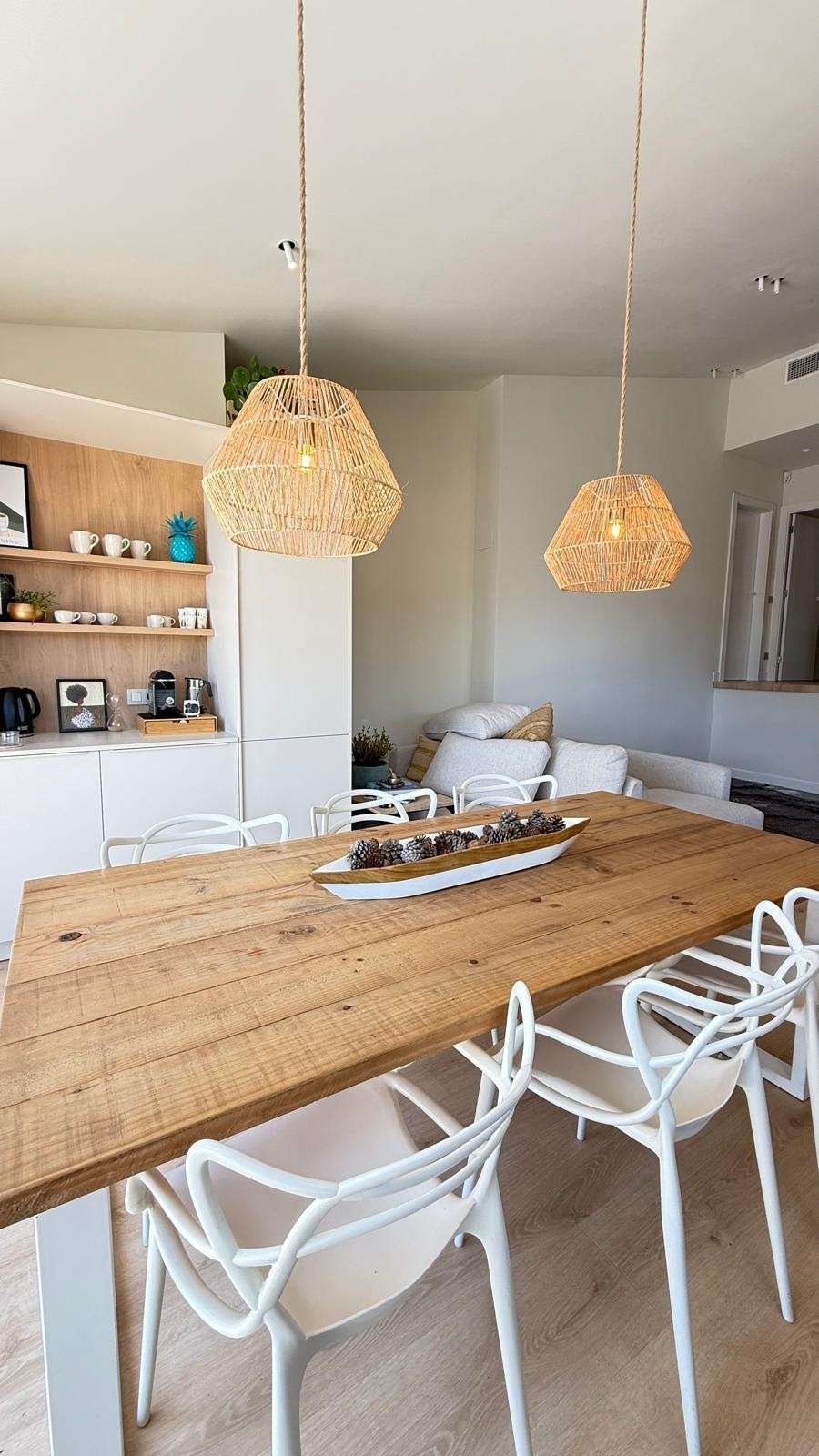 Comedor luminoso con mesa de madera, sillas blancas, lámparas colgantes tejidas y un sofá gris al fondo.